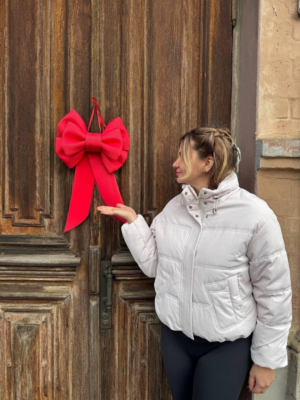 May include: A large, bright red bow hanging on a weathered wooden door. The bow is the focal point, with long, flowing ribbons. The door has vertical panels and a dark metal handle. The background includes a building.