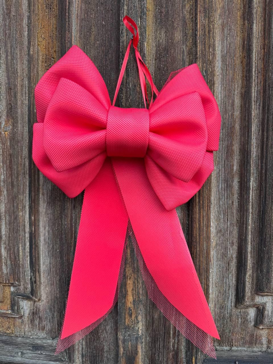 May include: A large, vibrant pink bow with long, flowing tails hangs against a weathered wooden background. The bow is made of a textured fabric, and the tails have a sheer, mesh-like trim. A red ribbon is used for hanging.