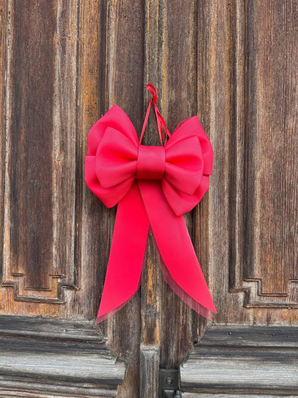 May include: A vibrant, hot pink bow with long, flowing tails hangs against a weathered, wooden door. The bow is made of a textured fabric, and the tails have a delicate, sheer trim. The bow is suspended by a thin, red loop.