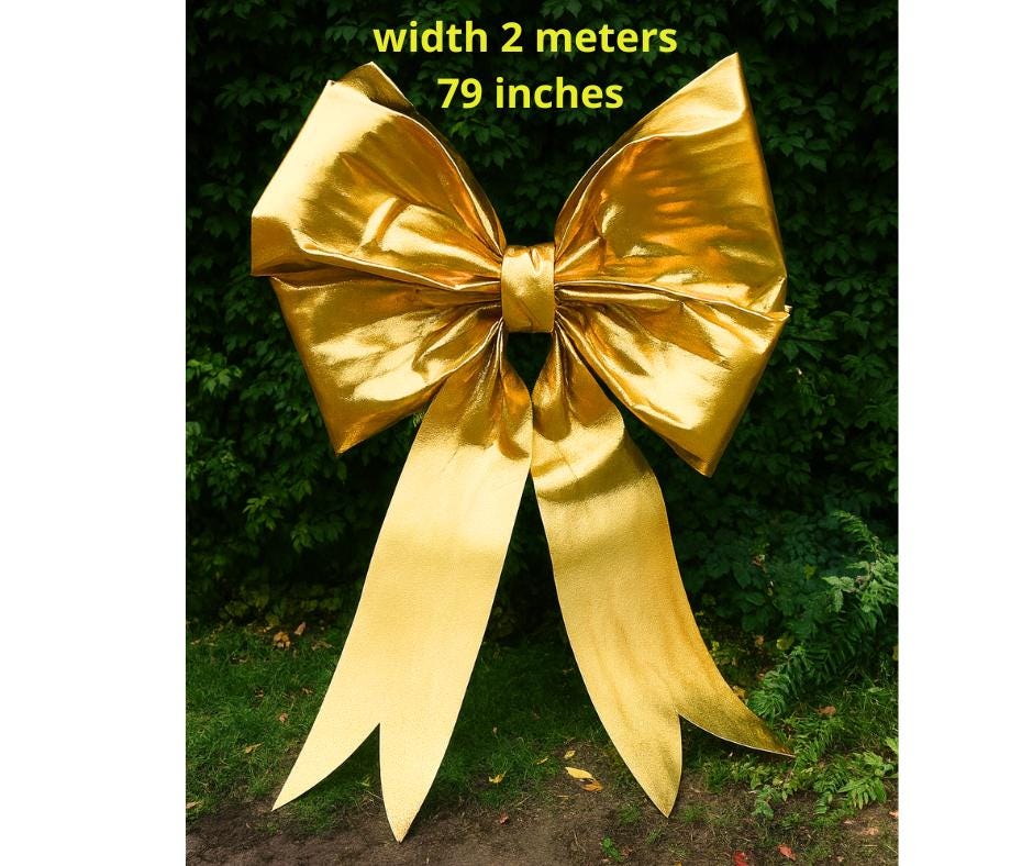 May include: A large, golden bow with long, flowing ribbons. The bow is approximately 79 inches wide and is set against a backdrop of green foliage. The bow's metallic sheen catches the light, creating a festive and eye-catching display.