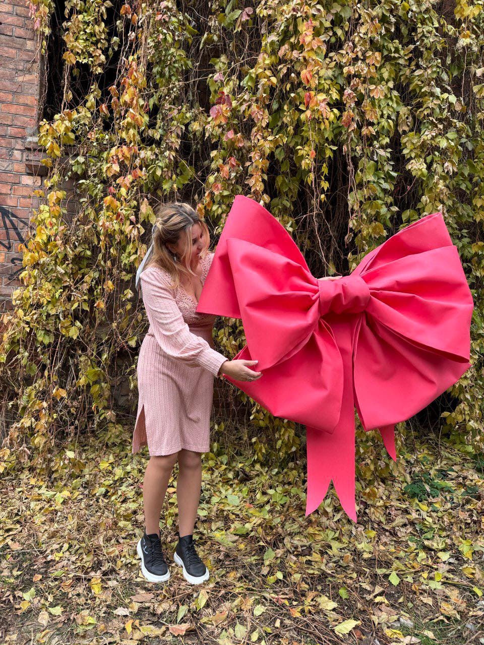 May include: A large, vibrant pink bow, possibly for decoration or a gift. The bow is held by a person in a pink dress and black sneakers, set against a backdrop of autumn foliage and a brick wall.
