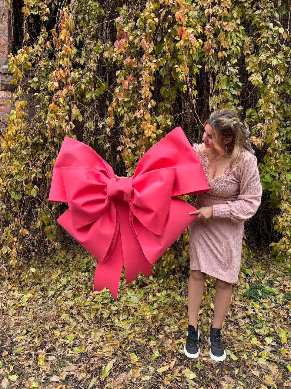 May include: A large, vibrant pink bow, likely for decoration, is held up in front of a background of autumn foliage. The bow is the focal point, with its multiple loops and tails. The bow's color contrasts with the natural setting.