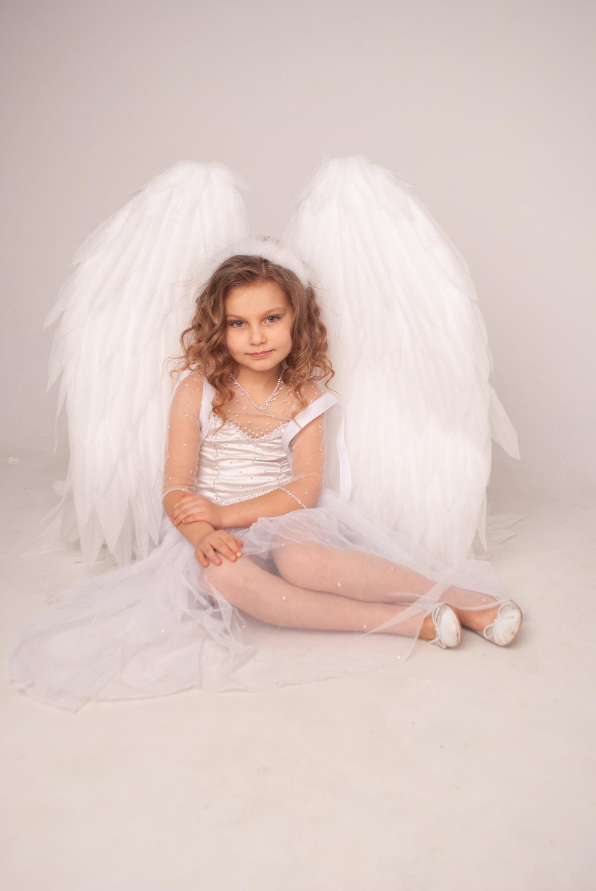 May include: A young person in a white angel costume, including large feathered wings, a dress, and a halo. The person is seated, with arms crossed, and wearing white shoes. The outfit is embellished with sequins.