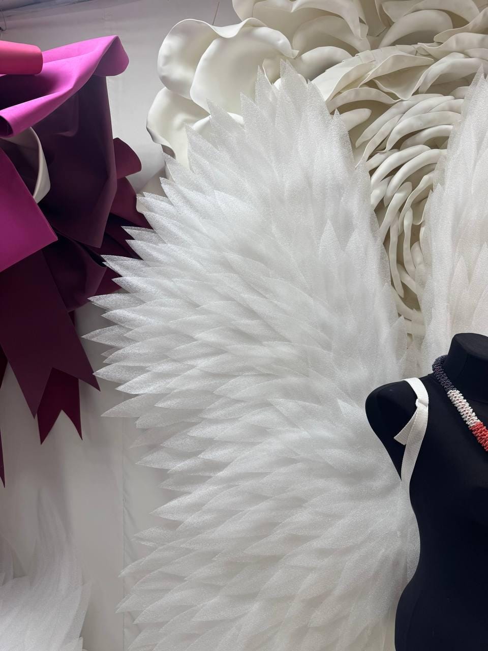 May include: A display featuring large, white, feathery wings, possibly for a costume or art installation. The wings are made of a textured, layered material. A black mannequin torso with a necklace and white straps is visible.