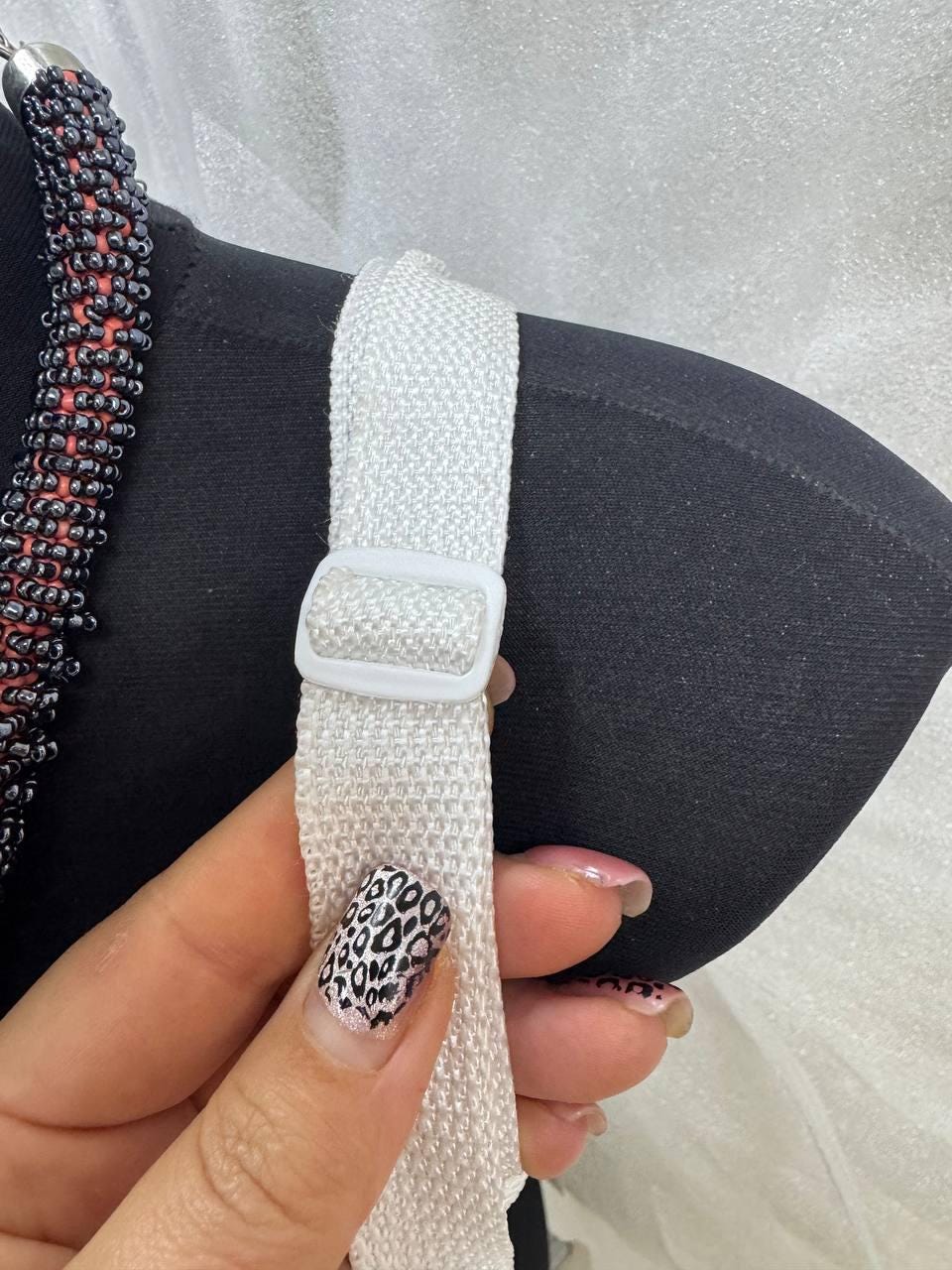 May include: Close-up of a black bra with a white adjustable strap and a white plastic slider. A beaded necklace with black and red beads is visible on the left. The image includes a hand with a leopard print nail design.