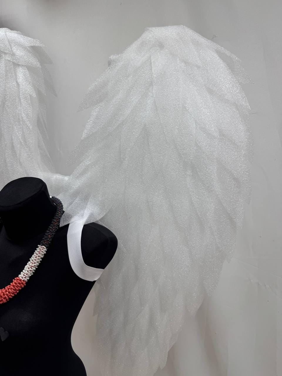 May include: White angel wings made of a textured material, displayed behind a black mannequin torso. A colorful beaded necklace with red, white, and black beads is draped on the mannequin.