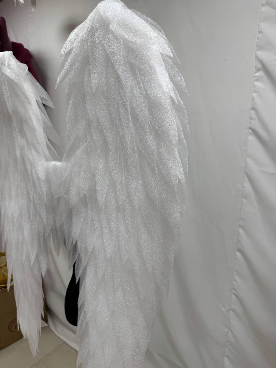 May include: A pair of large, white angel wings with a layered, feathery design. The wings are made of a textured, possibly foam-like material, and are displayed against a white backdrop. The wings are designed for costume or decorative purposes.