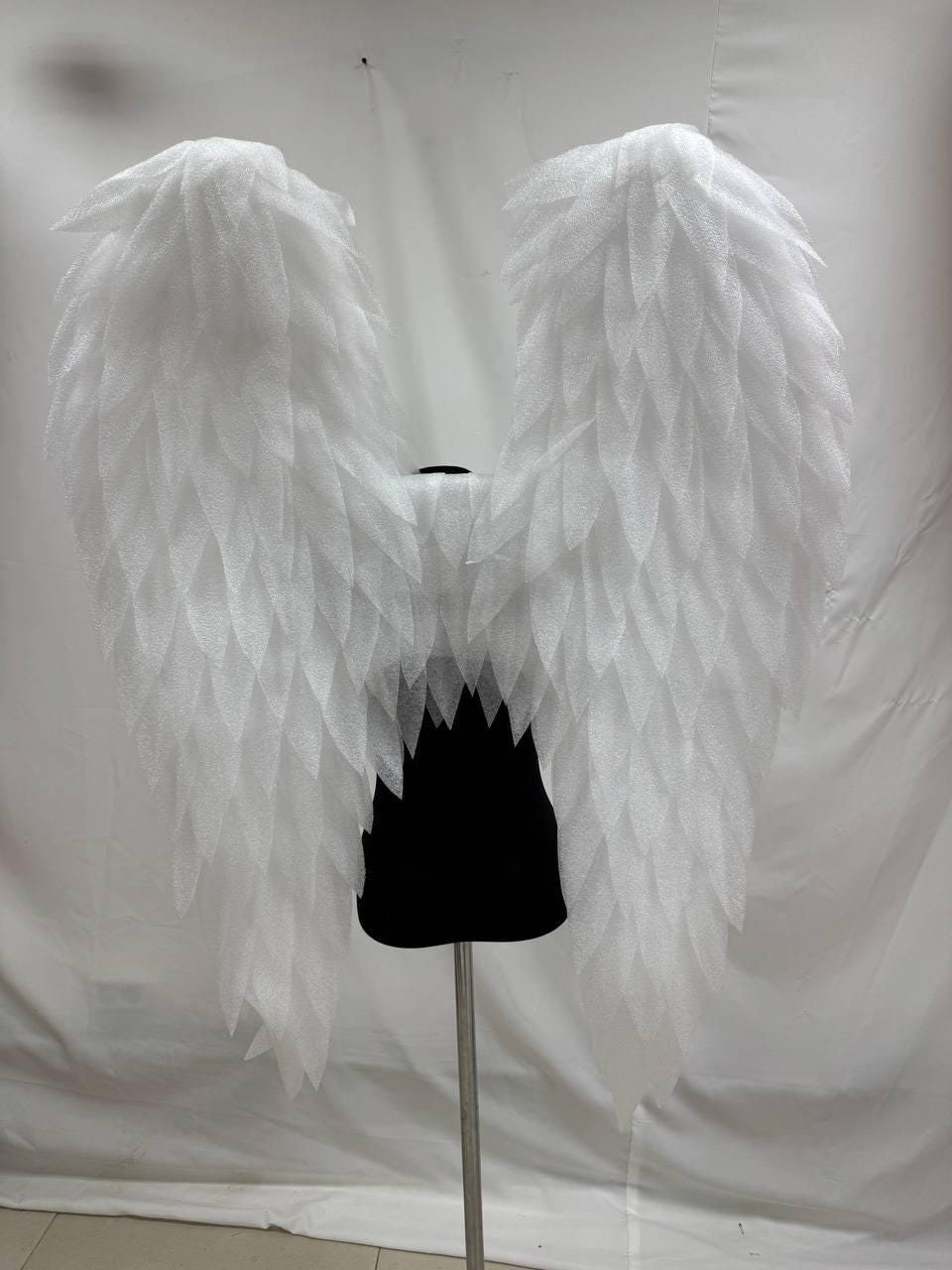 May include: Large, white angel wings with a feathery texture. The wings are displayed on a black form, attached to a silver stand. The wings are symmetrical and appear to be made of a lightweight material.