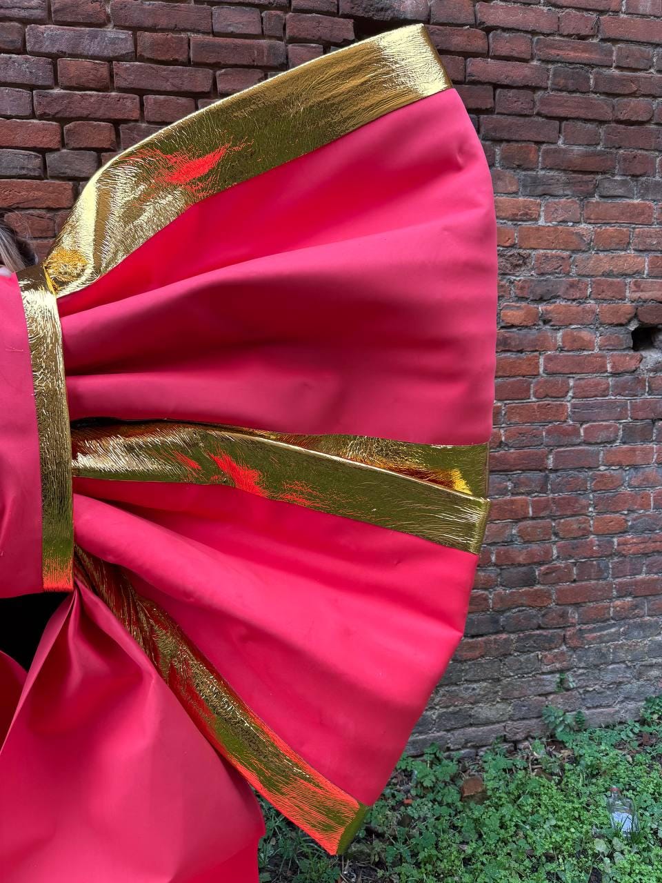May include: A large, vibrant pink bow with gold trim. The bow is made of a smooth, shiny fabric and is set against a brick wall background. The gold trim adds a touch of elegance to the design.