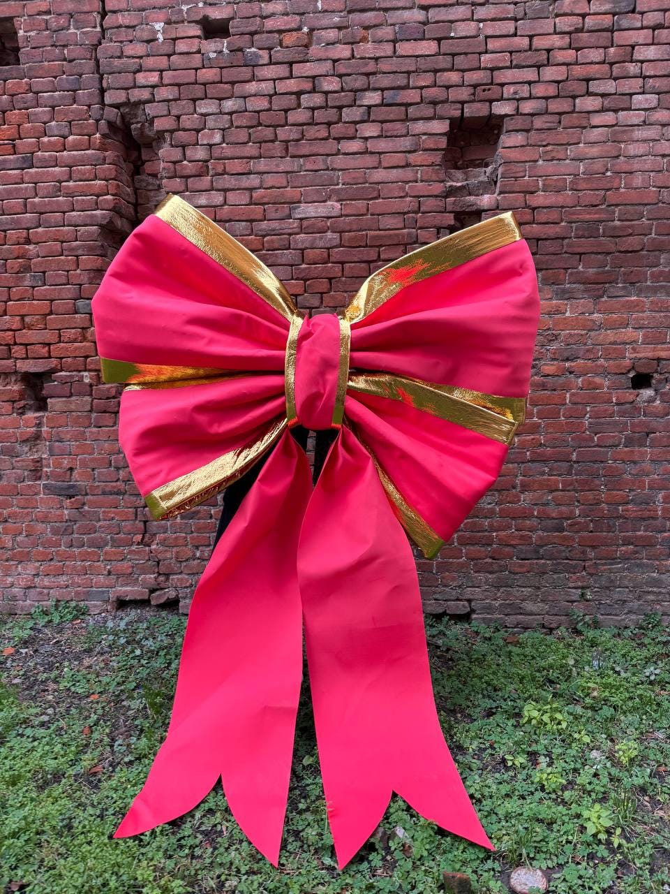 May include: A large, vibrant red bow with gold trim. The bow is made of a shiny fabric and has long, flowing tails. The background is a weathered brick wall and green foliage, creating a festive and eye-catching display.