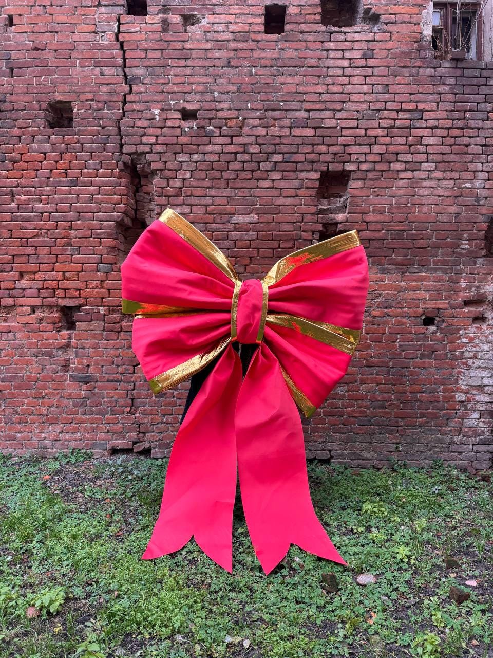 May include: A large, vibrant red bow with gold trim. The bow is made of a shiny material and has long, flowing tails. The background is a weathered brick wall and green grass.