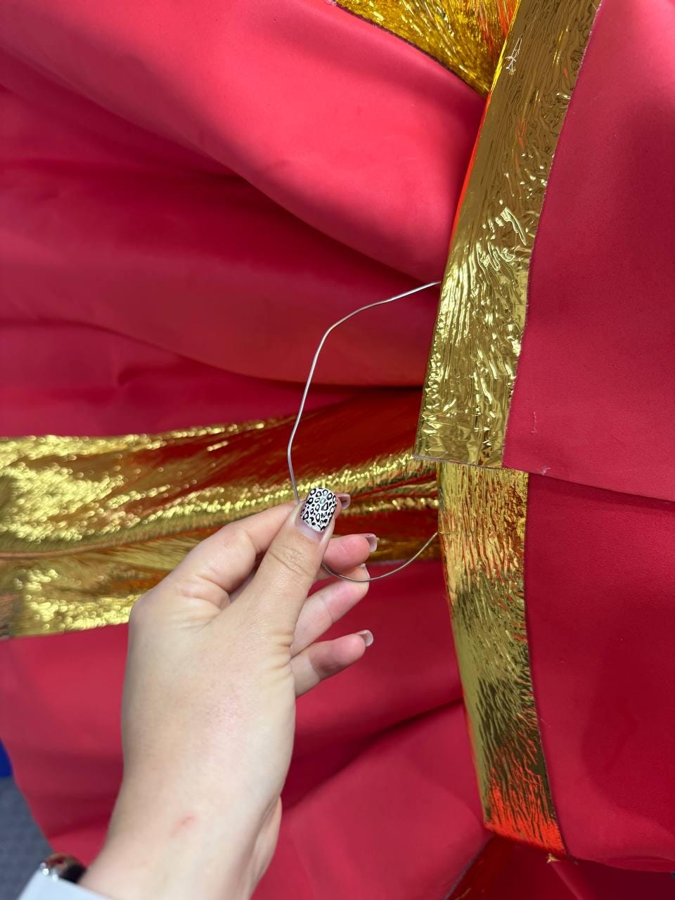 May include: Close-up of a large, red fabric bow with gold trim. A hand with a patterned nail design holds a silver wire, possibly for securing the bow. The bow's folds and metallic accents create a festive, decorative appearance.