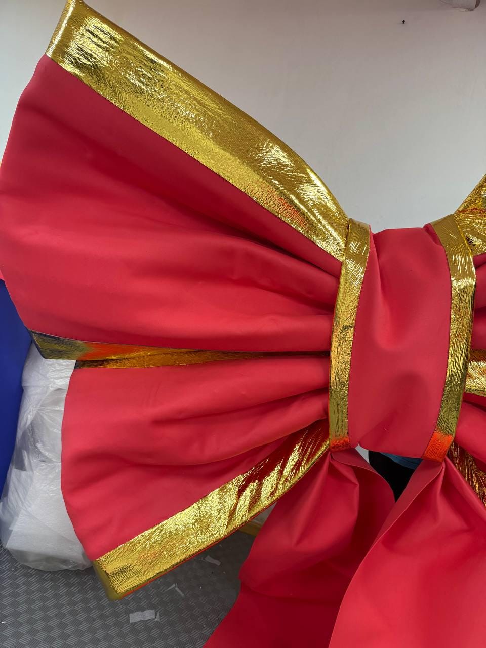 May include: A large, decorative bow in a vibrant red fabric with gold trim. The bow's edges and center band are accented with shiny gold material, creating a festive and eye-catching design. This oversized bow is suitable for holiday decorations or special events.