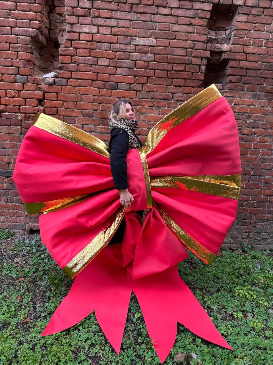 May include: A large, bright red bow with gold trim. The bow is oversized and appears to be made of fabric. The background is a brick wall and green foliage.