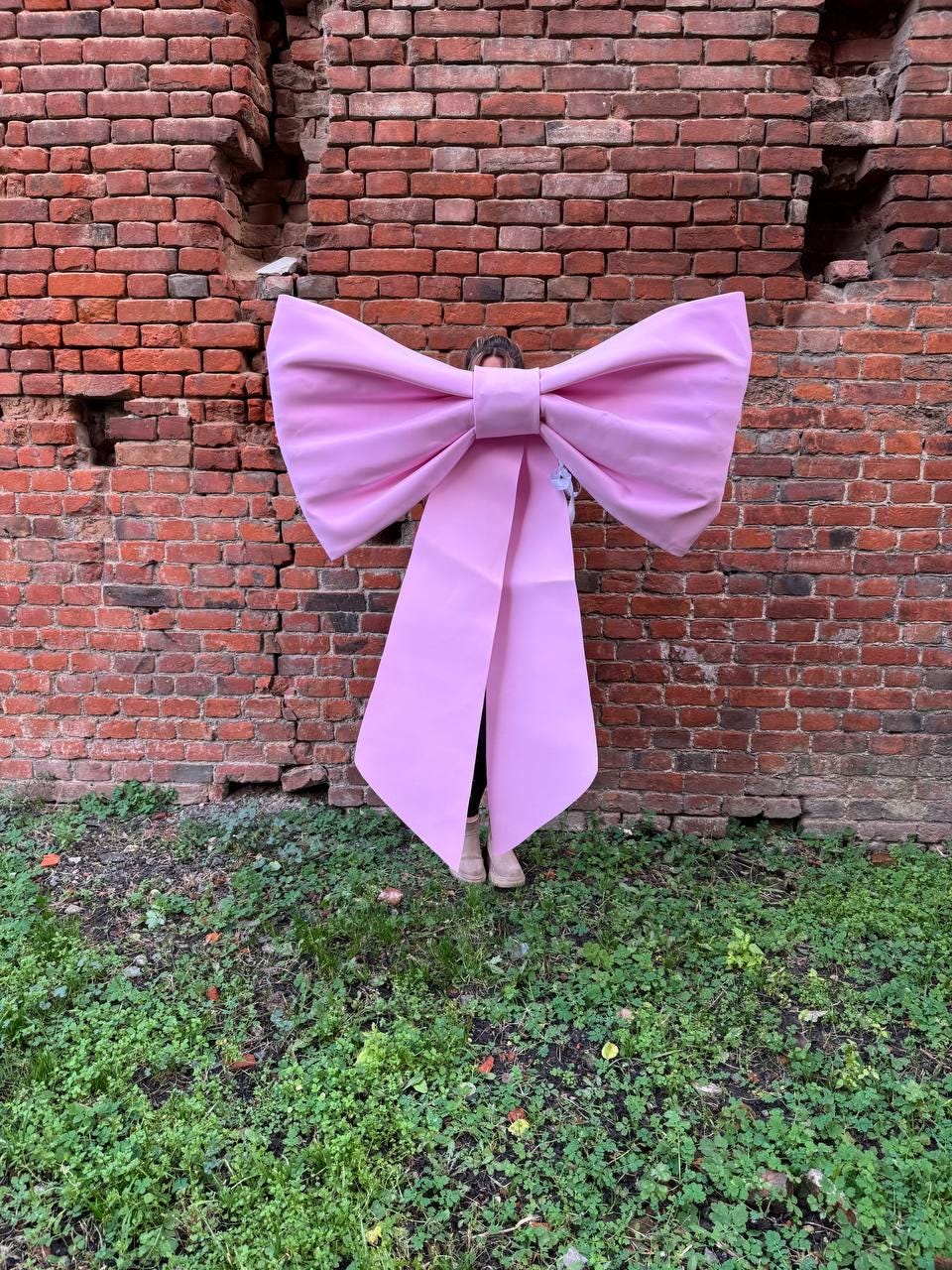 May include: A large, light pink bow prop with long, flowing tails. The bow is held in front of a person, against a backdrop of a weathered brick wall and green foliage. The bow's size suggests it's a costume or decorative element.