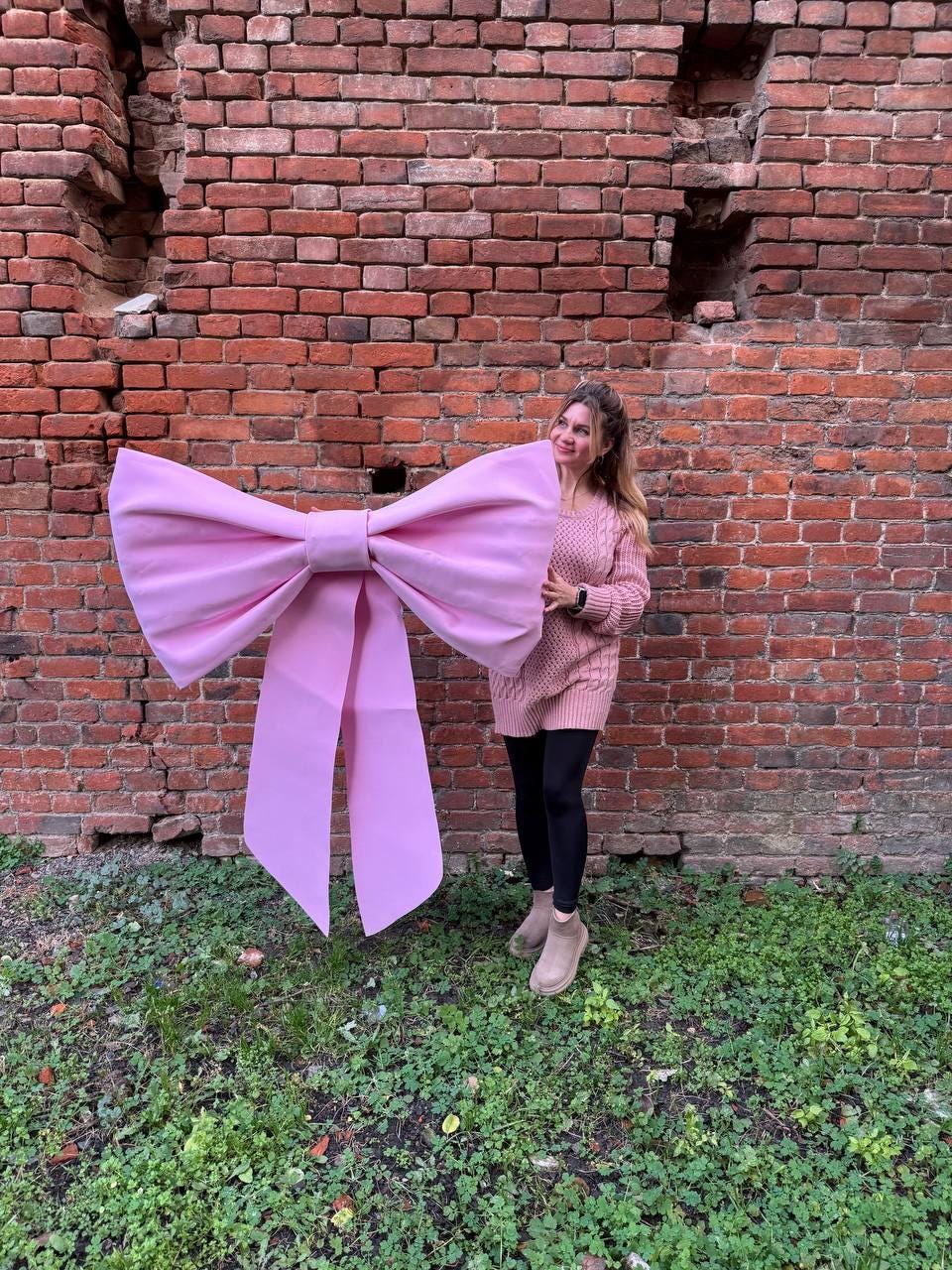 May include: A large, light pink bow with long streamers. The bow is held in front of a brick wall. The bow is a solid color and appears to be made of a sturdy material. The background includes green foliage.