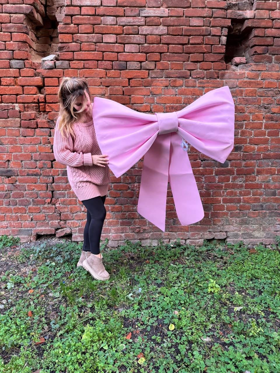 May include: A large, light pink bow with long streamers. The bow is held in front of a brick wall. The bow is a decorative item, possibly for a party or event.