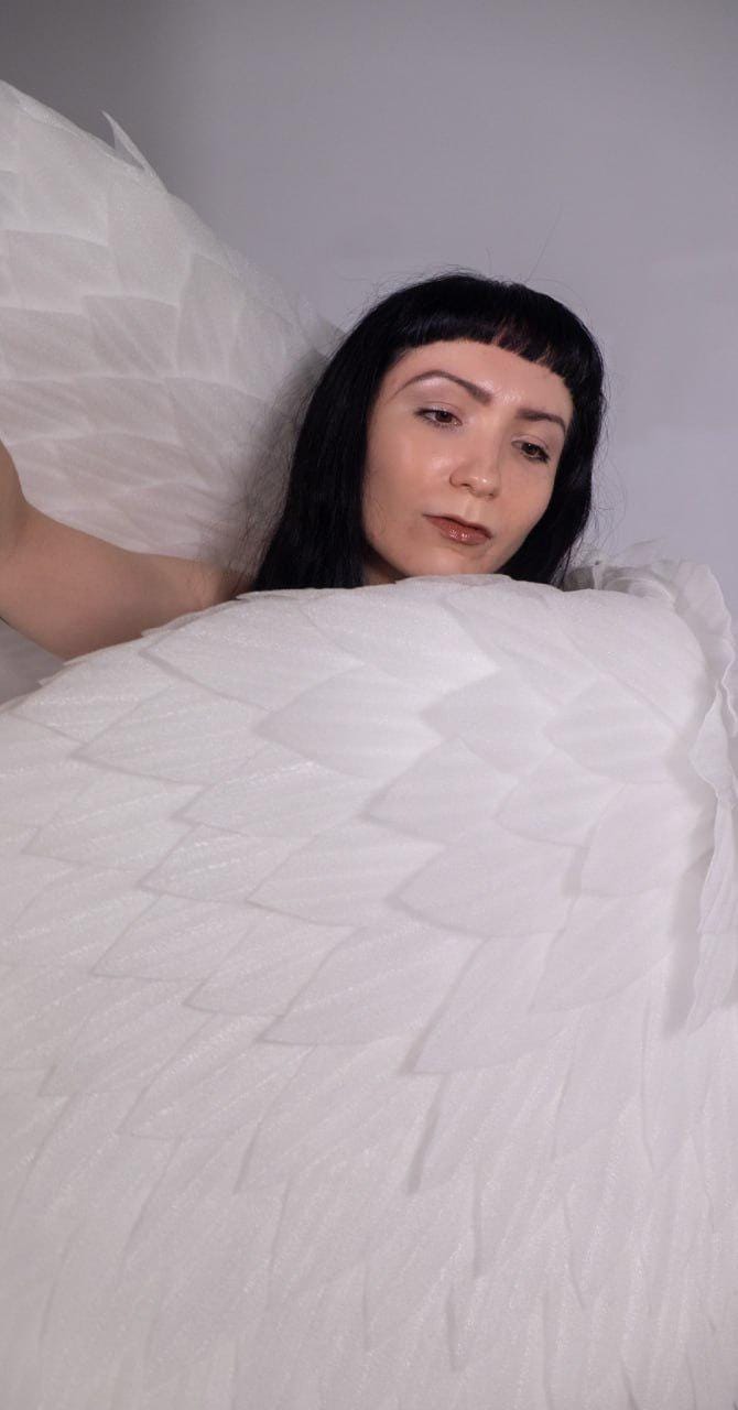 May include: A person with long black hair and bangs is partially visible behind a large, white, feathery object. The object has a layered, textured appearance, resembling wings or a decorative textile.