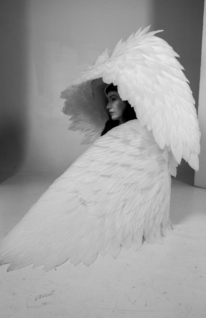 May include: A black and white image of a person wearing large, white feathered wings. The wings are detailed and expansive, creating a dramatic effect. The person is seen in profile, with dark hair and a serene expression. The overall aesthetic is ethereal and artistic.