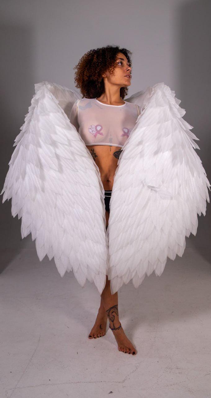 May include: A person wearing large, white angel wings. The wings are made of layered, textured feathers. The person is wearing a sheer, white top with pink ribbon designs. The background is a plain, light gray.