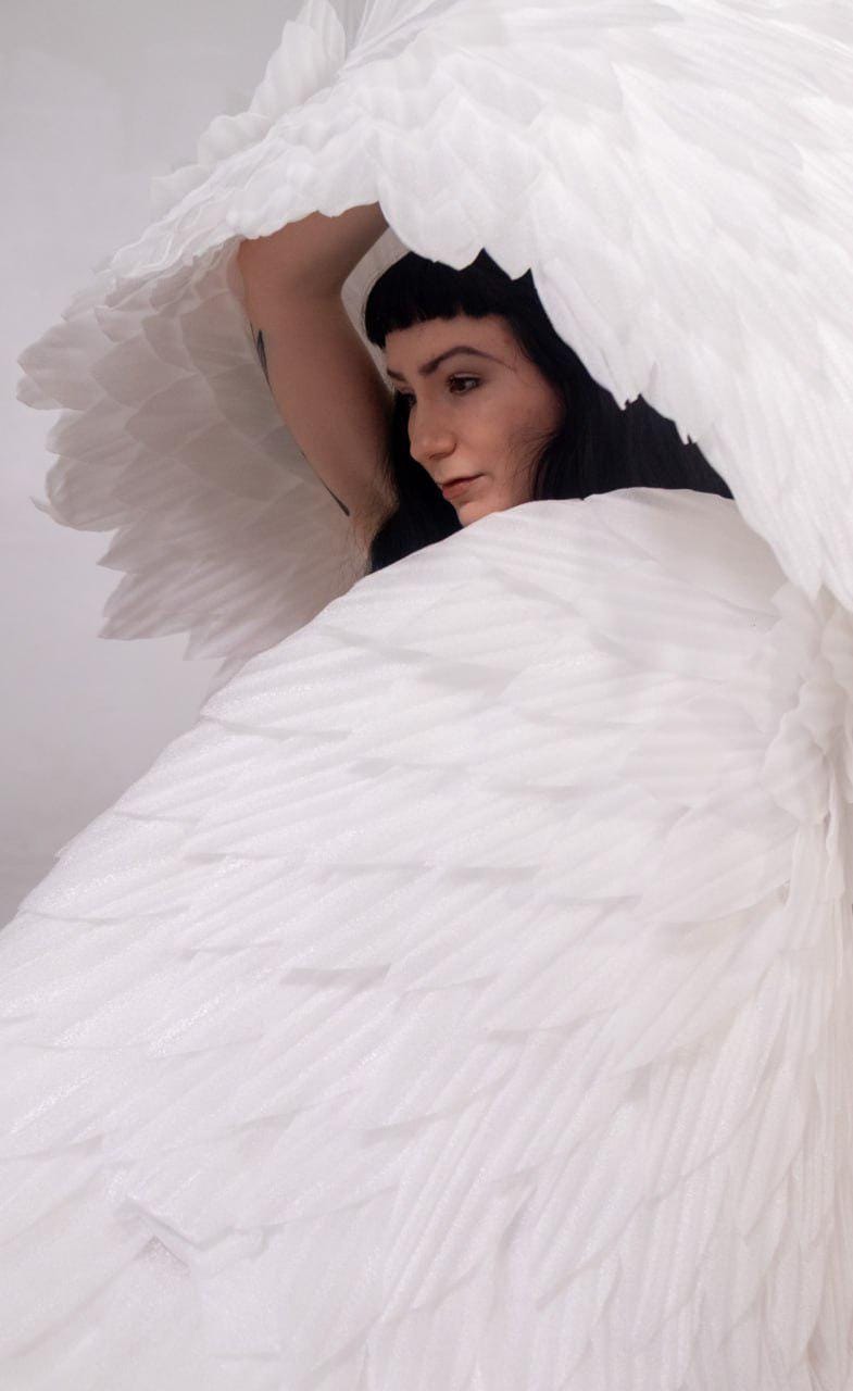 May include: A person is partially visible, framed by large, white, layered wings. The wings have a feathery texture and create a sense of movement. The person has dark hair and is looking down.