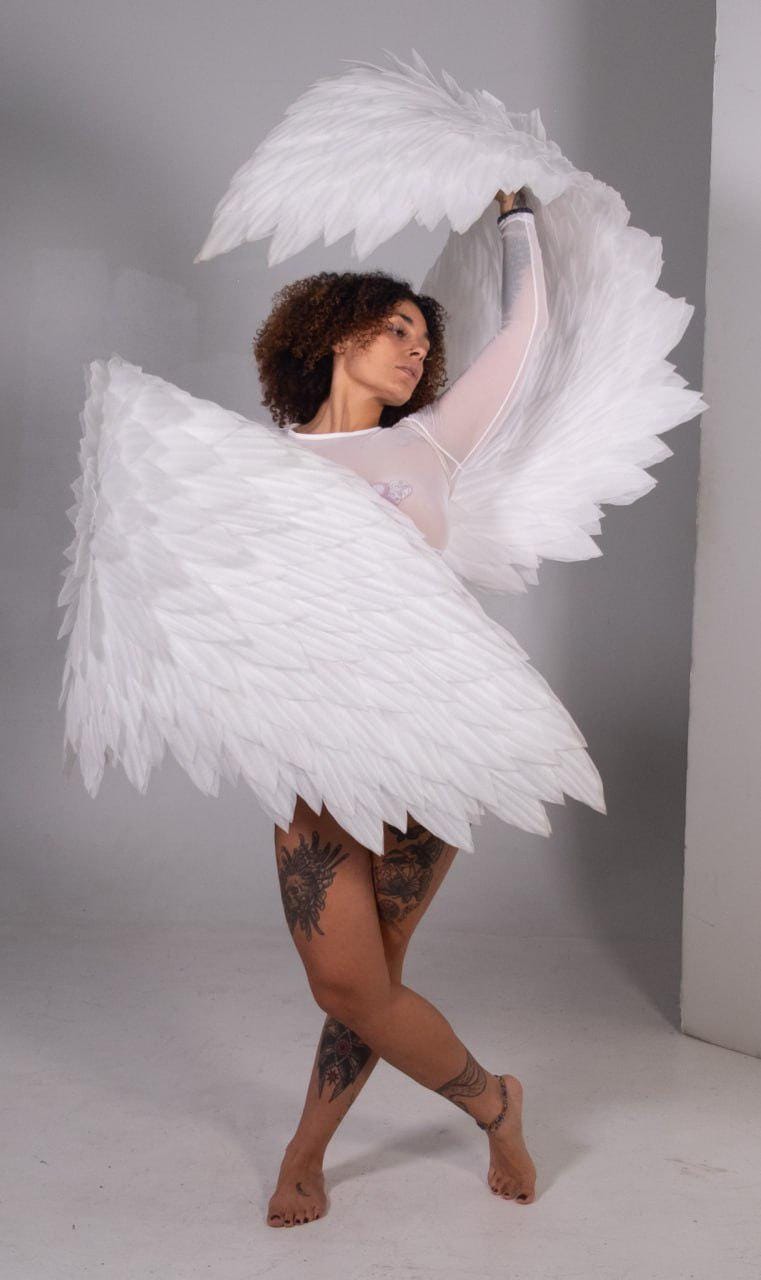 May include: A person wearing a sheer white top and large white feathered angel wings. The wings are spread wide, creating a dramatic pose. The person has dark curly hair and tattoos on their legs.