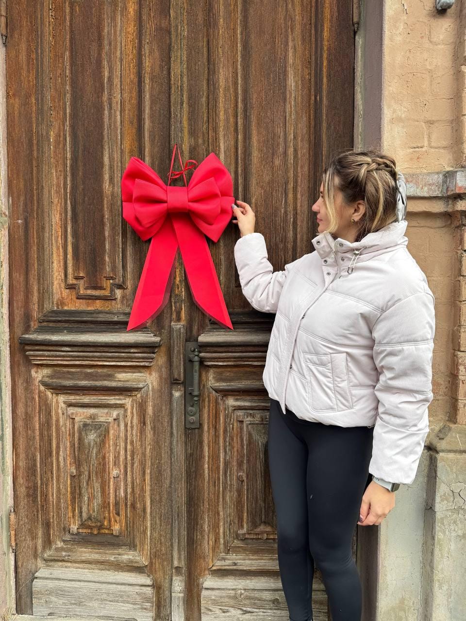 May include: A large, bright red bow is hanging on a weathered wooden door. The bow is the focal point, with long, flowing ribbons. The door has a dark, aged appearance, with visible wood grain and paneling. The bow adds a festive touch.