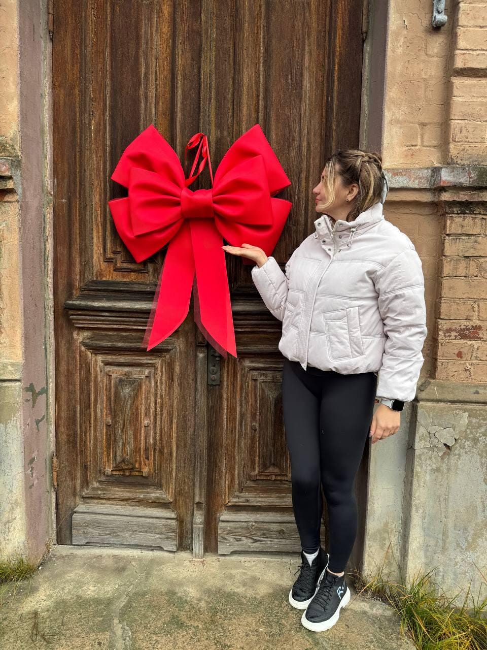 May include: A large, vibrant red bow hangs on a weathered wooden door. The bow is made of a soft material and has long, flowing tails. The door is a rich brown color and has intricate paneling. The bow is a festive decoration.