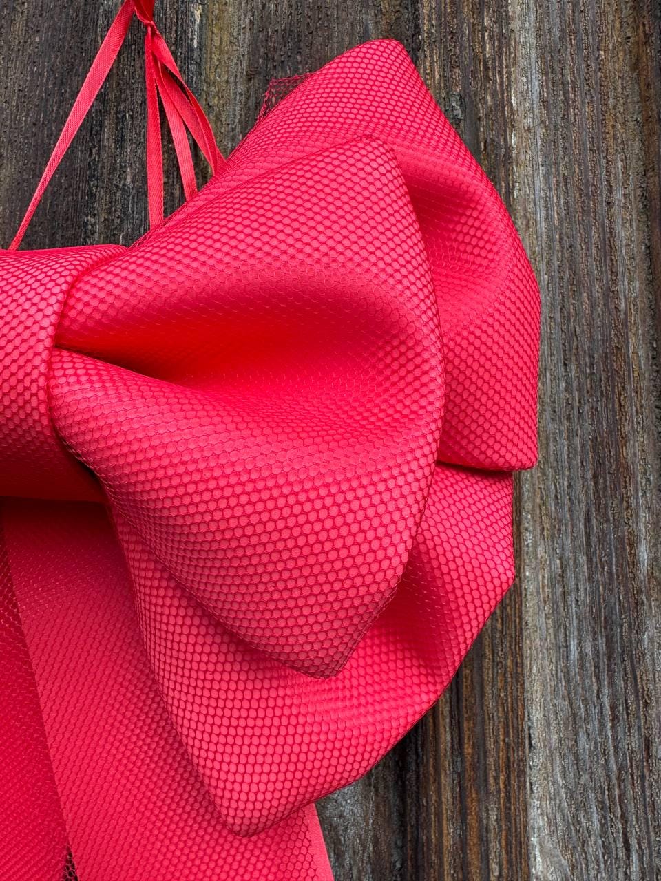 May include: A large, vibrant red bow with a textured, dotted pattern. The bow is made of a silky material and is tied with matching red ribbons. The background is a weathered wooden surface.