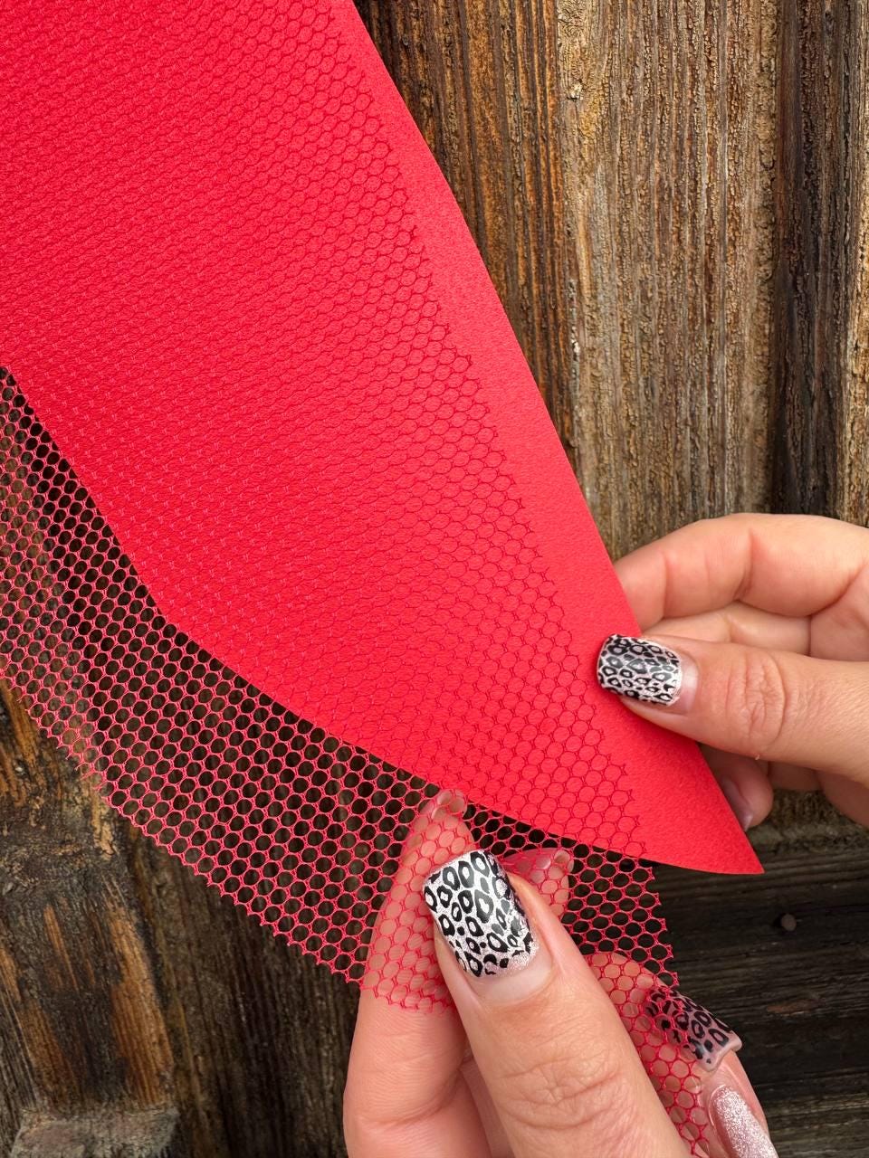 May include: A close-up of red fabric and red mesh material. The mesh has a hexagonal pattern. The fabric is held by a person with leopard print nail art. The background is a wooden surface.