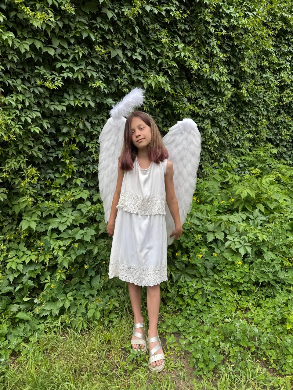 May include: A young person wearing a white dress with lace detailing, silver sandals, and large white angel wings. A white halo sits atop their head. The background is a lush green wall of foliage.