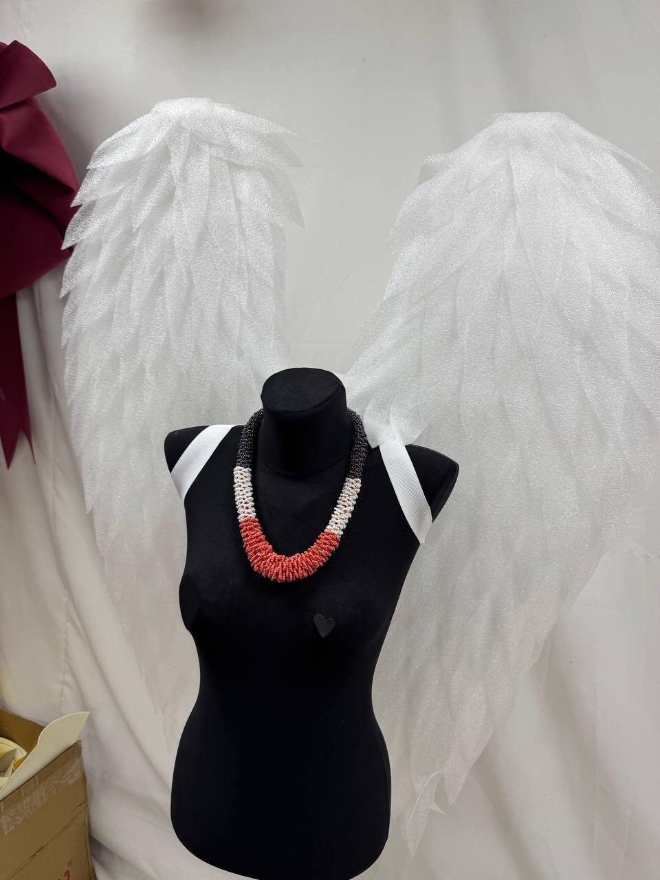 May include: A beaded necklace with black, white, and red sections is displayed on a black mannequin. The necklace is the focal point, with white angel wings in the background. The mannequin is wearing white straps.