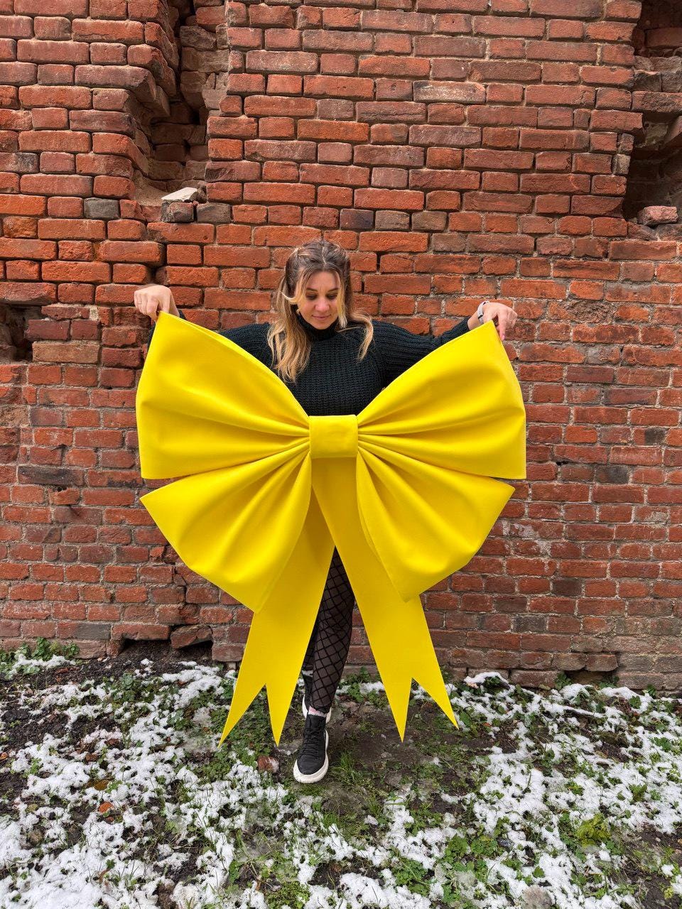 May include: A large, bright yellow bow with long, pointed tails. The bow is held up in front of a brick wall. The bow is made of a soft, possibly fabric material. The background includes a brick wall and patches of snow on the ground.