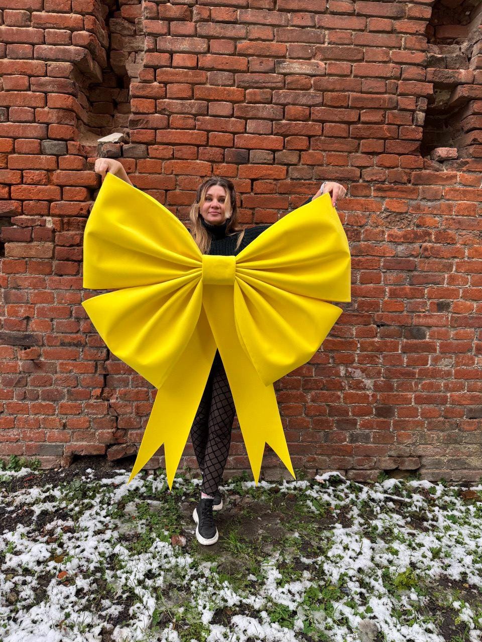 May include: A large, bright yellow bow with long tails. The bow is held in front of a brick wall. The bow is a vibrant, eye-catching decoration, perfect for events or as a statement piece.