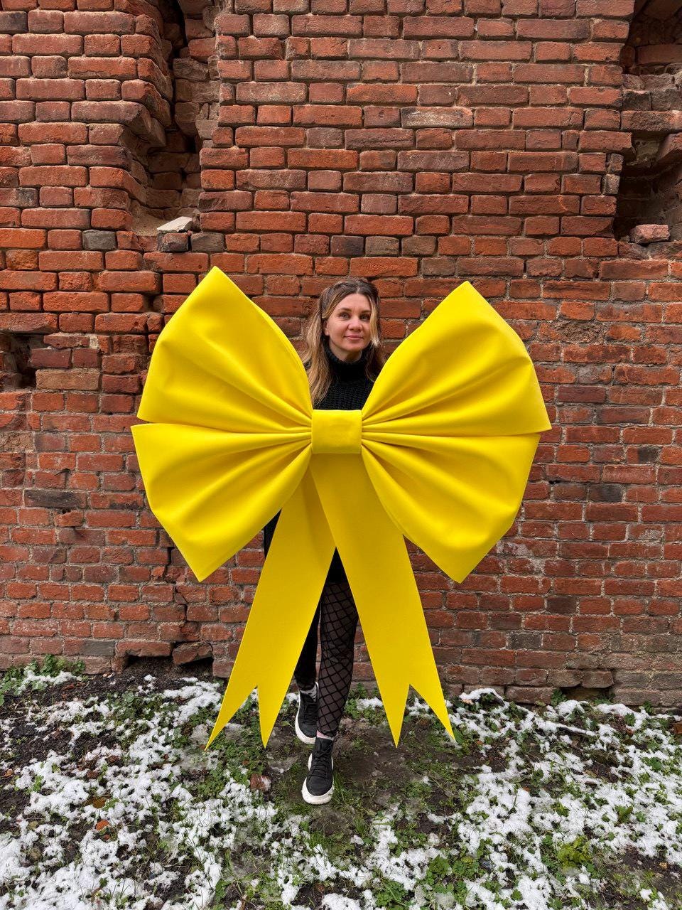 May include: A large, bright yellow bow with long, pointed streamers. The bow is held in front of a person standing in front of a brick wall. The bow is a vibrant, eye-catching decoration.