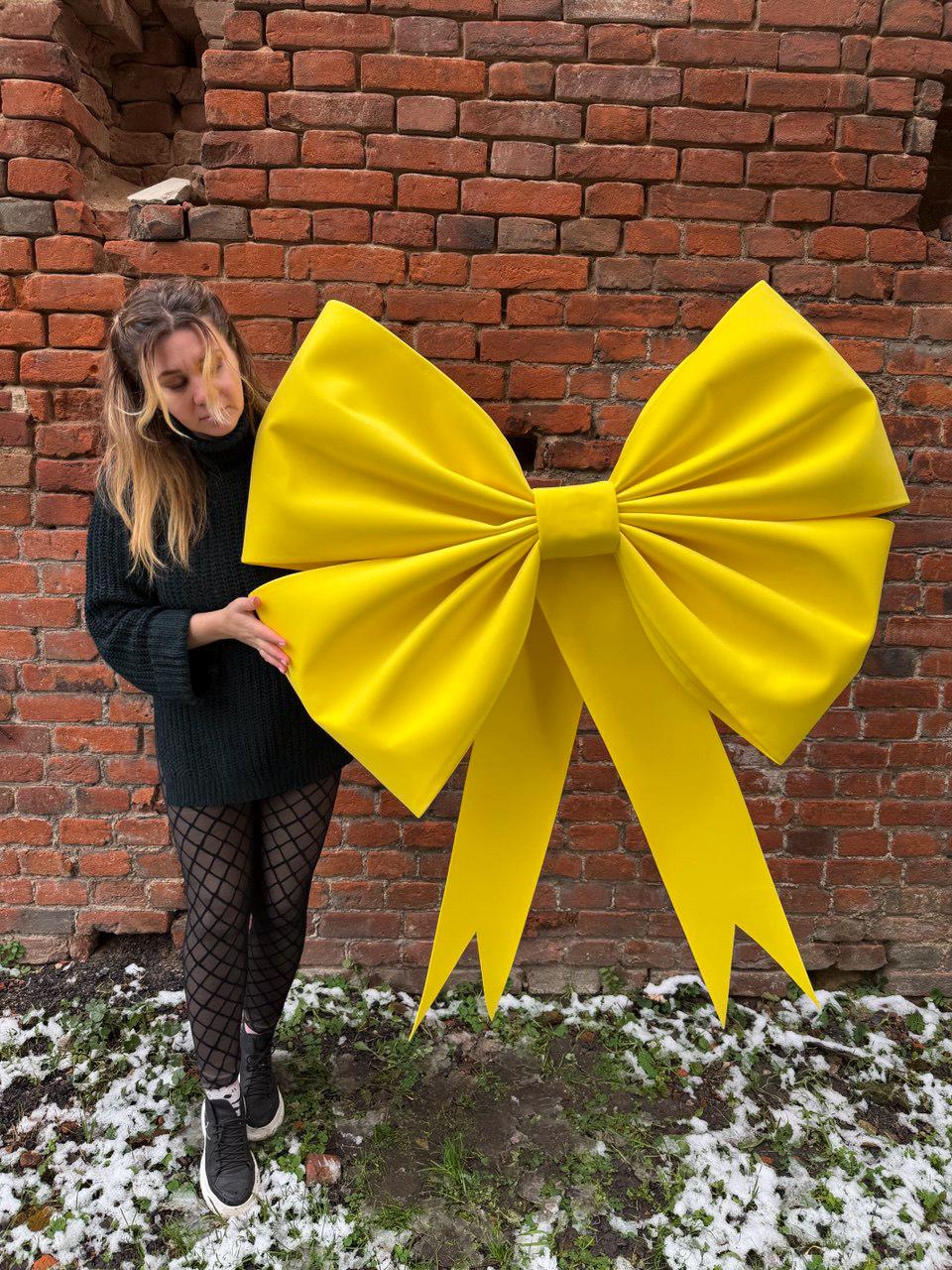 May include: A large, bright yellow bow with long, pointed streamers. The bow is made of a soft, possibly fabric material. The bow is displayed in front of a brick wall.