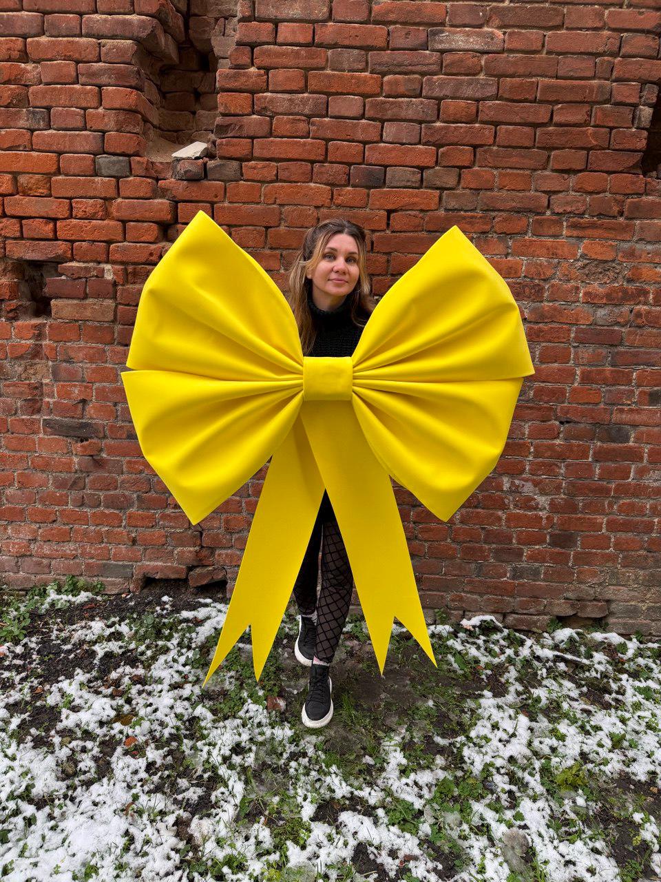 May include: A large, bright yellow bow with long, flowing tails. The bow is made of a soft, velvety material and is positioned in front of a brick wall. The bow is a vibrant accent against the neutral background.