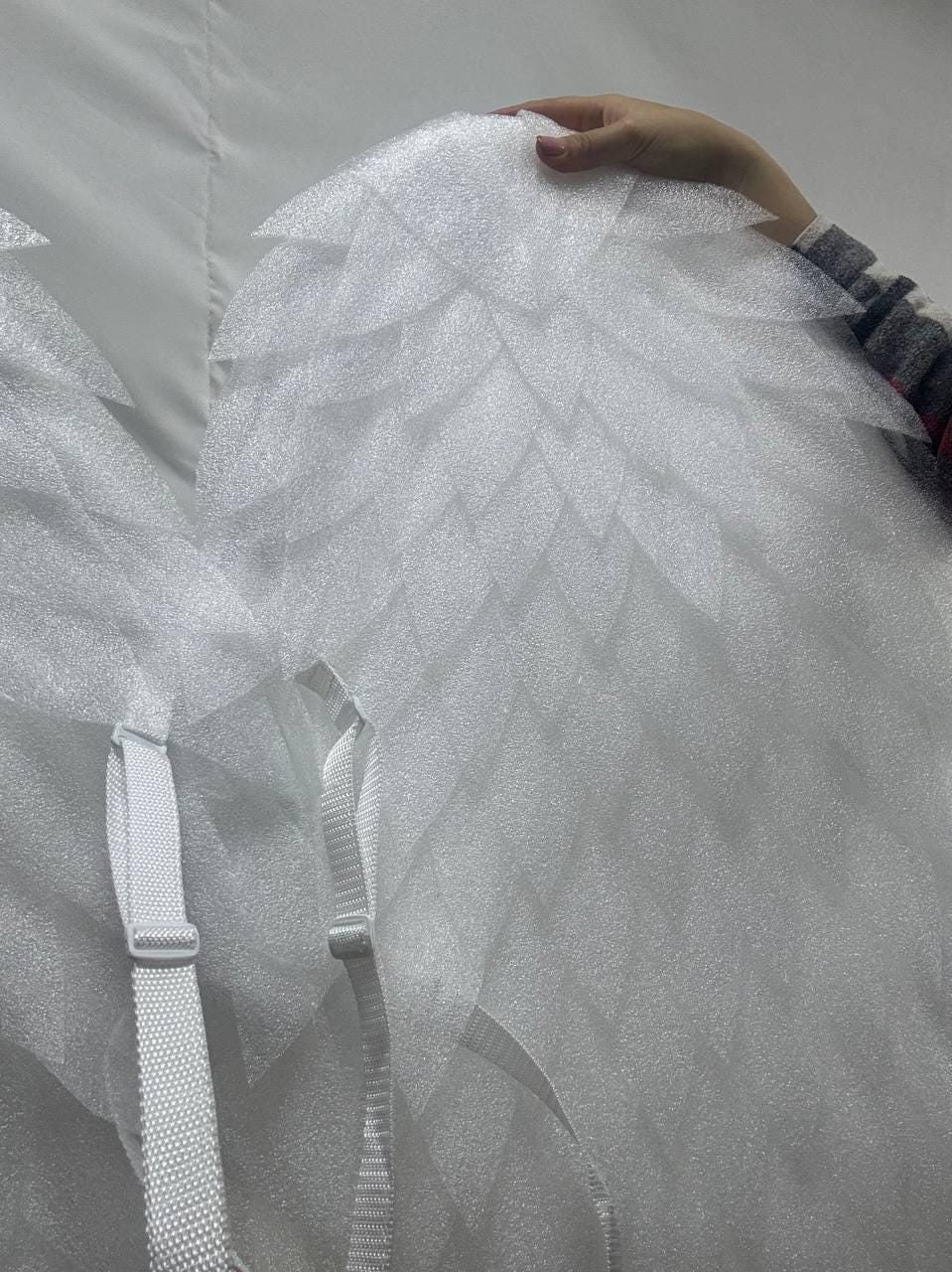 May include: White angel wings with a layered, textured design. The wings have a series of pointed, feather-like shapes. White adjustable straps are attached for wearing. The wings are held by a person's hand.