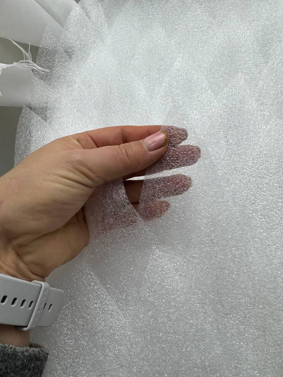 May include: A close-up of a hand holding a piece of white, textured foam packaging material. The foam has a bubbly, porous appearance. The background is filled with more of the same packaging material.