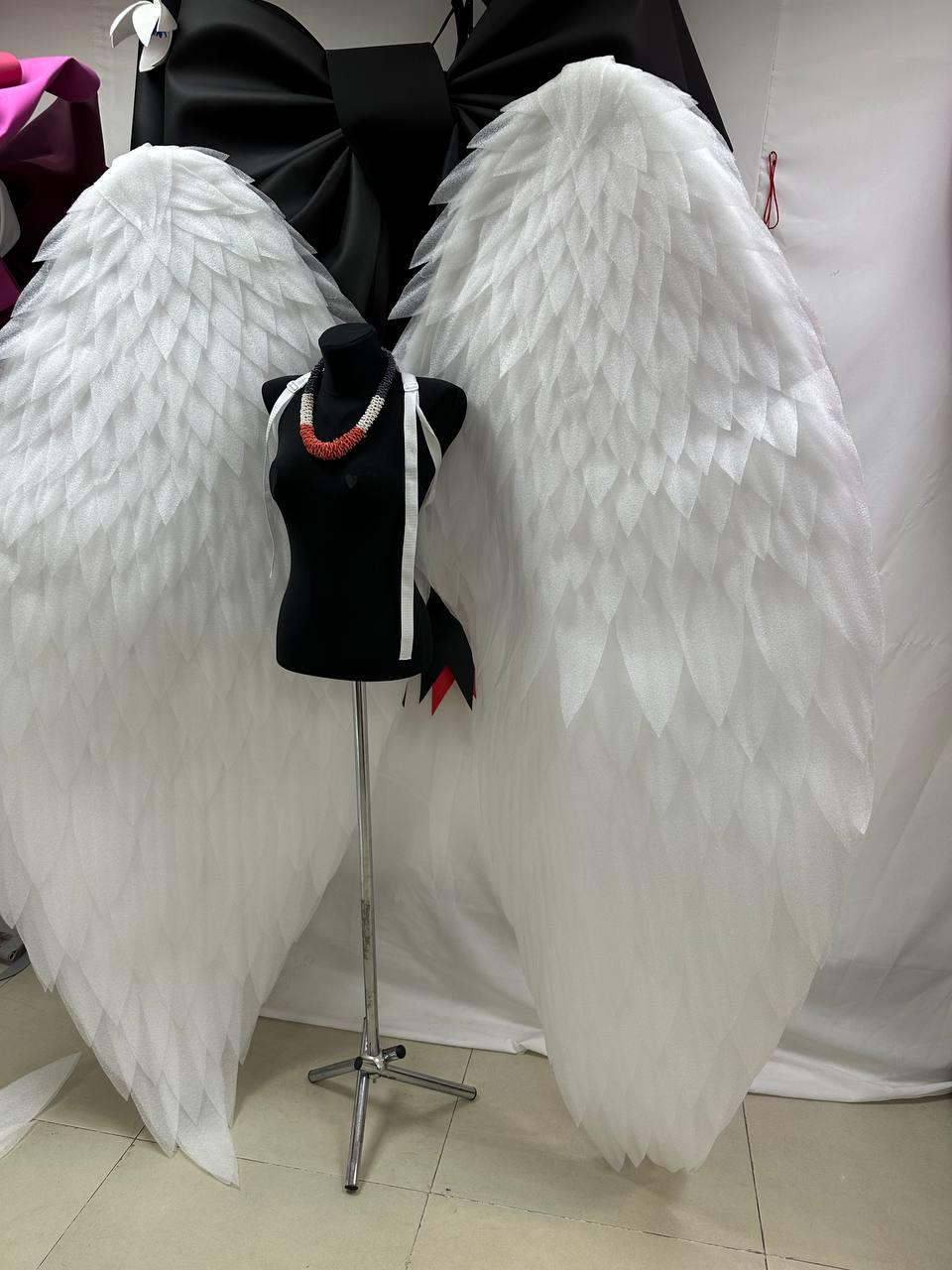 May include: Large white angel wings with a layered feather design. A black mannequin is displayed in front of the wings, wearing a necklace with red and white beads. A black bow is visible in the background.