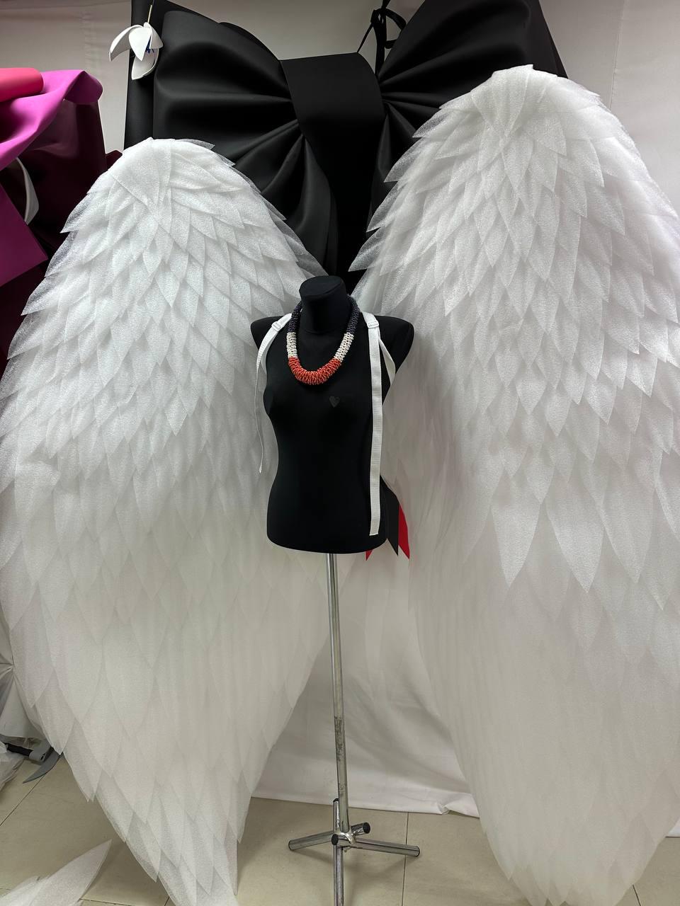 May include: A black mannequin is adorned with a white beaded necklace and white straps, positioned in front of large white angel wings. A large black bow is visible in the background. The wings have a layered, feathered appearance.