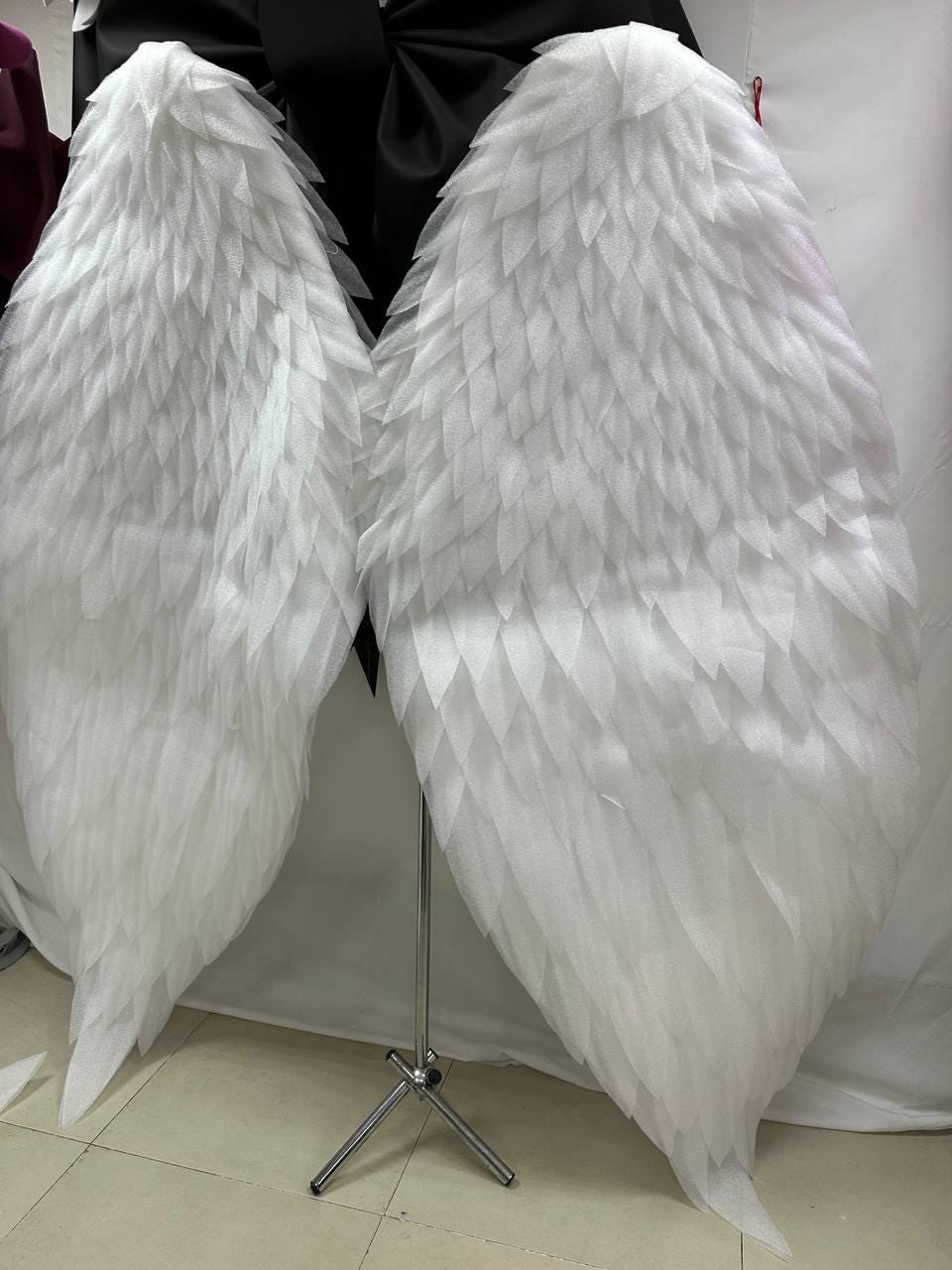 May include: Large, white angel wings with a layered feather design. The wings are displayed on a silver metal stand against a white background. The wings are made of a soft, textured material.