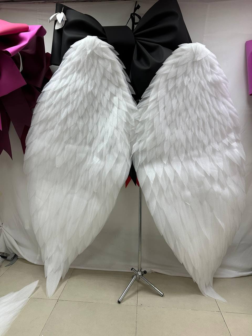 May include: A pair of large, white angel wings with a layered feather design. The wings are displayed on a silver stand against a white backdrop. A large black bow is visible in the background.
