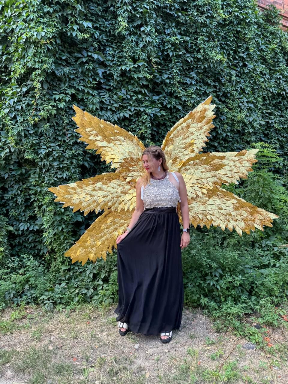 May include: A woman wearing a white and silver sequined top and a long black skirt stands in front of large golden angel wings. The wings are made of layered, textured material and are set against a backdrop of green foliage.