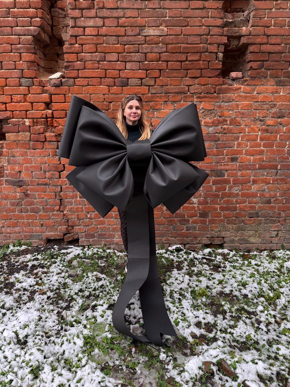 May include: A large, black bow with long, flowing tails. The bow is the focal point, set against a weathered brick wall and a patch of snow-covered ground. The bow's size and dark color create a striking visual contrast.