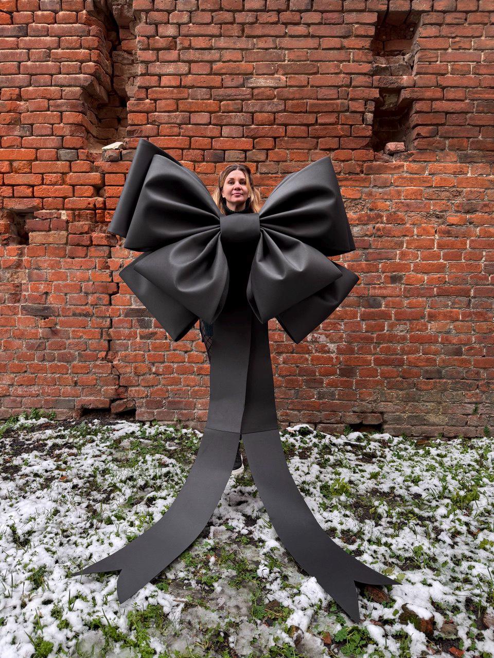 May include: A large, black bow with long, flowing tails. The bow is made of a matte material and is positioned in front of a brick wall. The bow is a striking decorative element, suitable for events or displays.