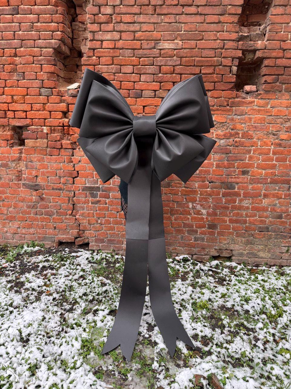 May include: A large, black bow with long, pointed streamers. The bow is made of a smooth, possibly satin-like material. It is set against a weathered brick wall with patches of snow and grass at the base.