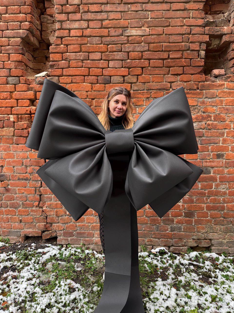 May include: A large, black bow made of a matte material. The bow is the focal point, with a woman standing behind it. The background is a weathered brick wall with patches of snow and greenery at the base.