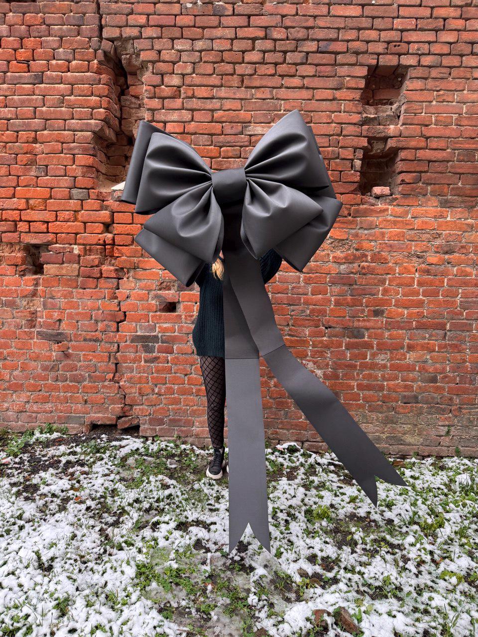 May include: A large, black decorative bow with long, flowing ribbons. The bow is made of a matte material and is held in front of a brick wall. The bow is a statement piece, perfect for events or decor.