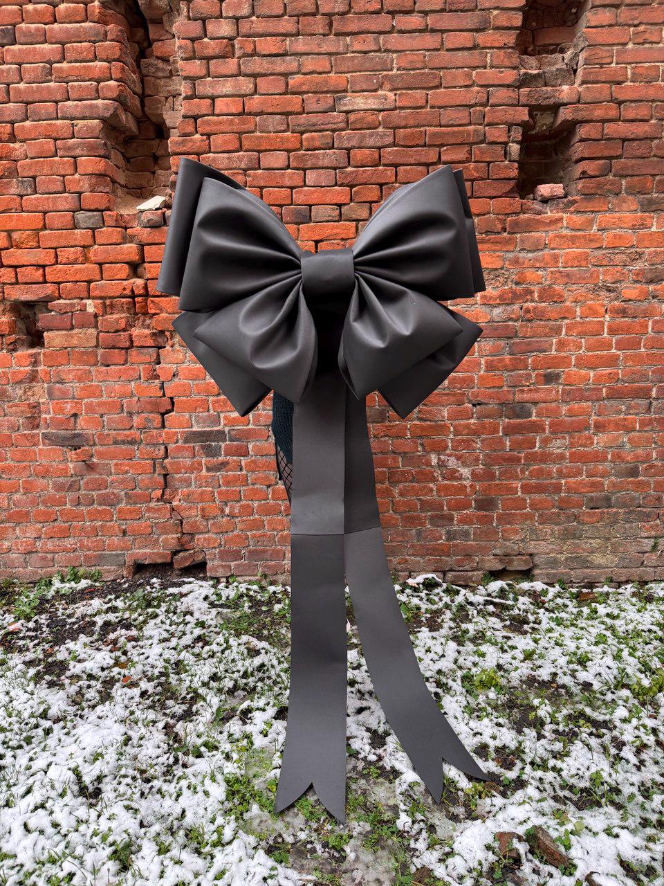 May include: A large, black decorative bow with long, flowing tails. The bow is made of a smooth, matte material and is set against a weathered brick wall and a patch of snow-covered ground. The bow's design is symmetrical and elegant.